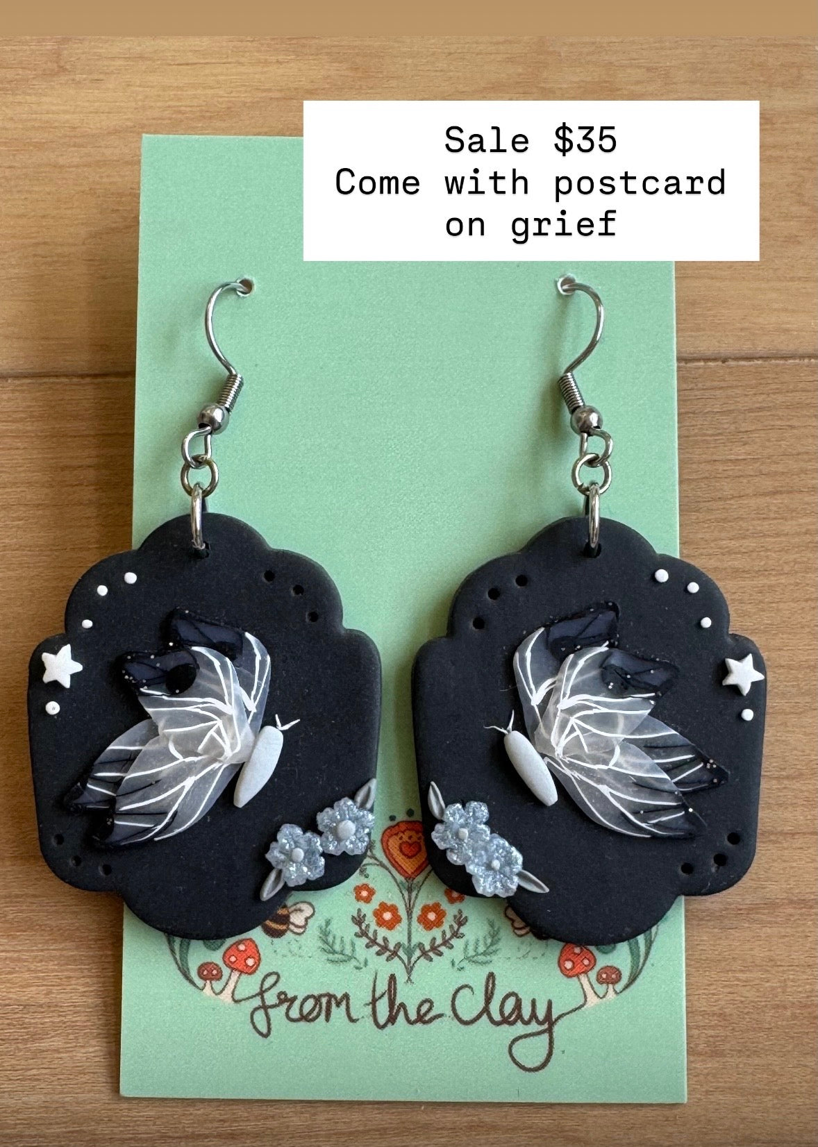 Black earrings with butterflies and small flowers on a green card with 'From the Clay' branding.