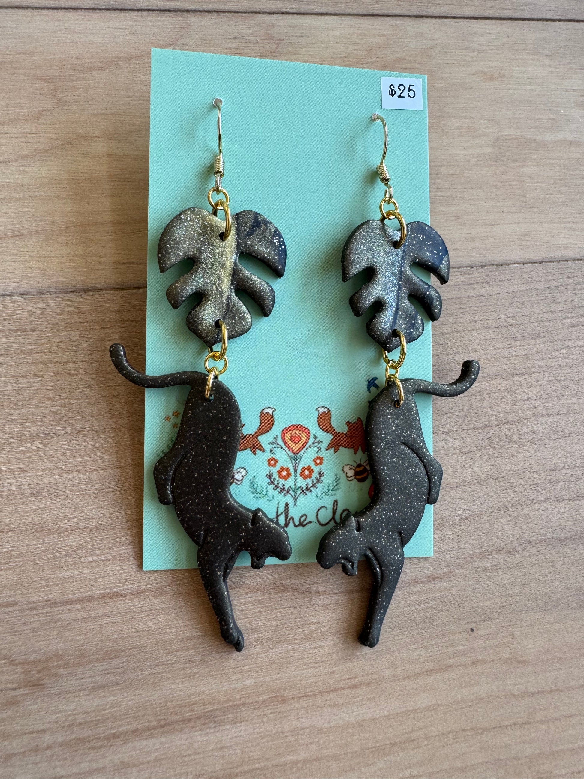 Pair of black and gold sparkly monstera leaf-shaped earrings with a black slightly sparkly panther attached. Placed  on a backing card on a wooden surface