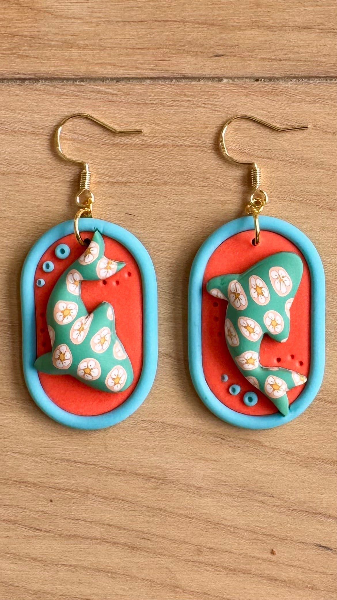 Pair of earrings with an orange oval background and a blue trim, with a floral design on a whale shark placed on a wooden surface