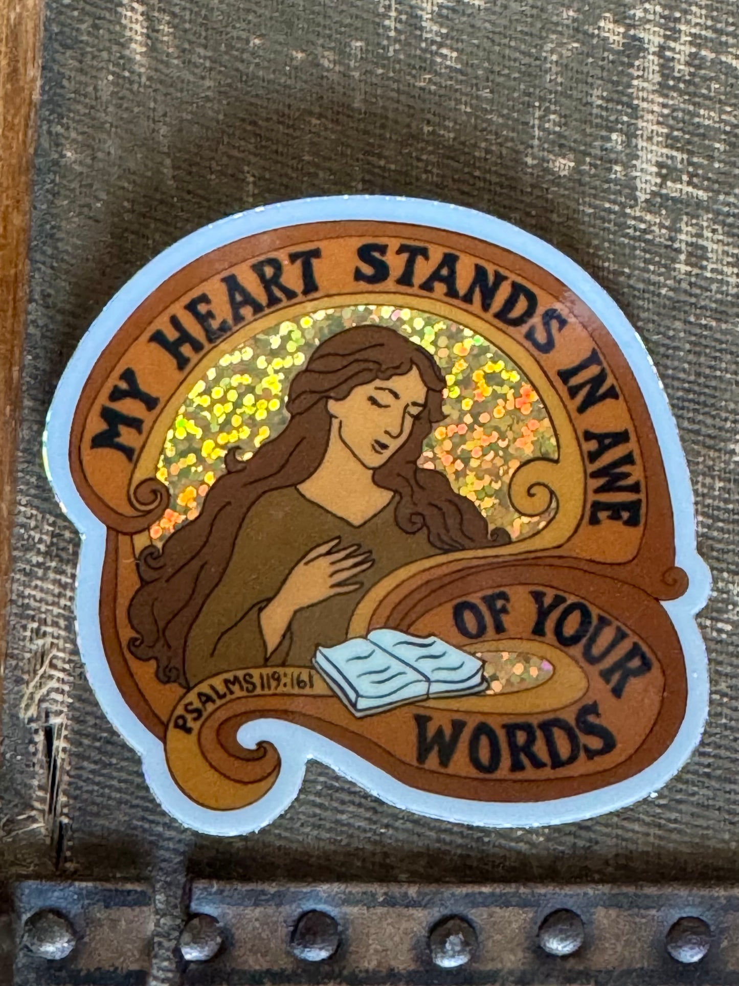 Sticker with a drawn woman holding a book and text 'My heart stands in awe of your words' the sticker is in brown and orange colors, with a glitter background behind the woman