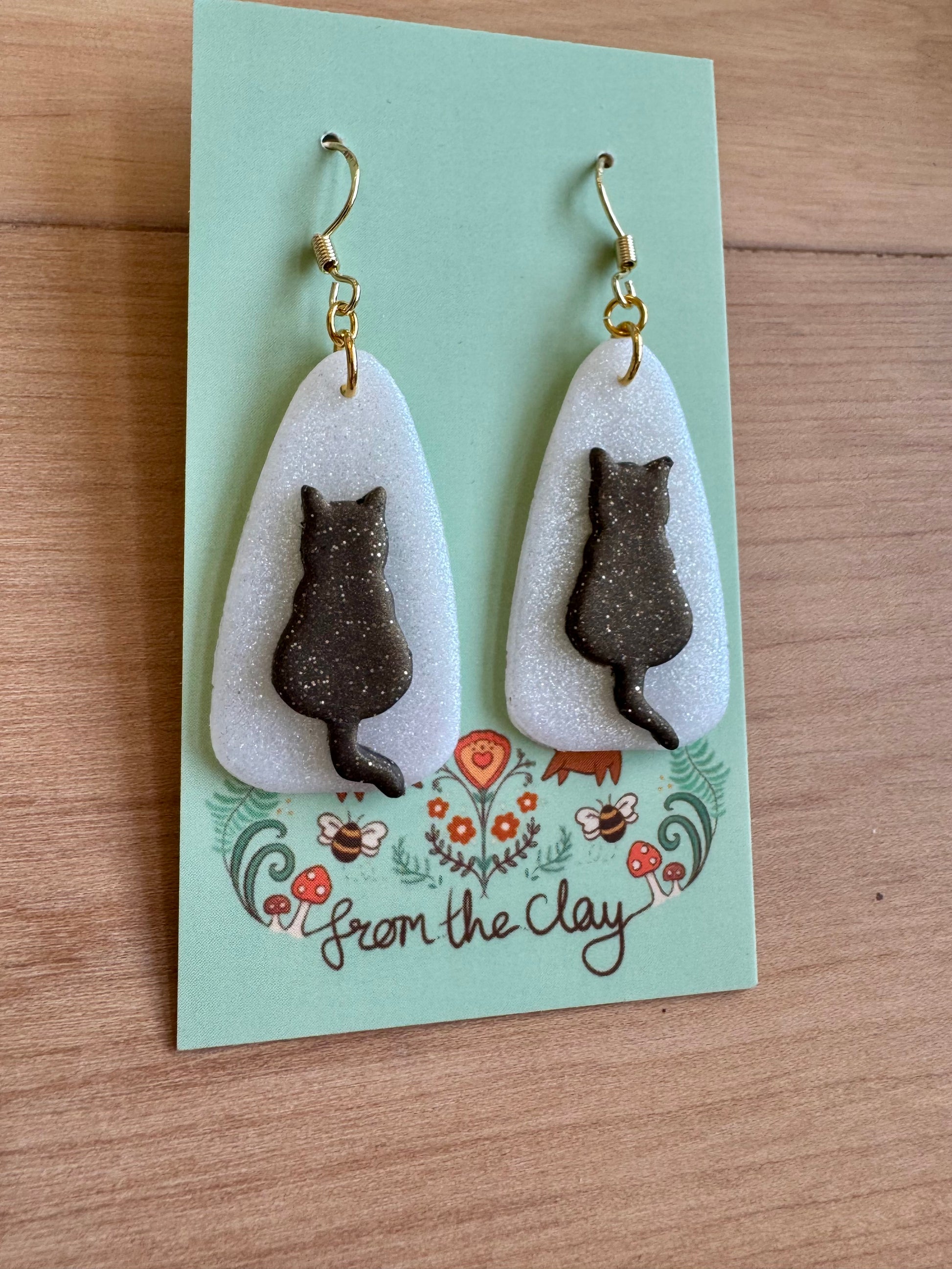 Black Cat silhouette with sparkle earrings on a 'From the Clay' branded card with a wooden background