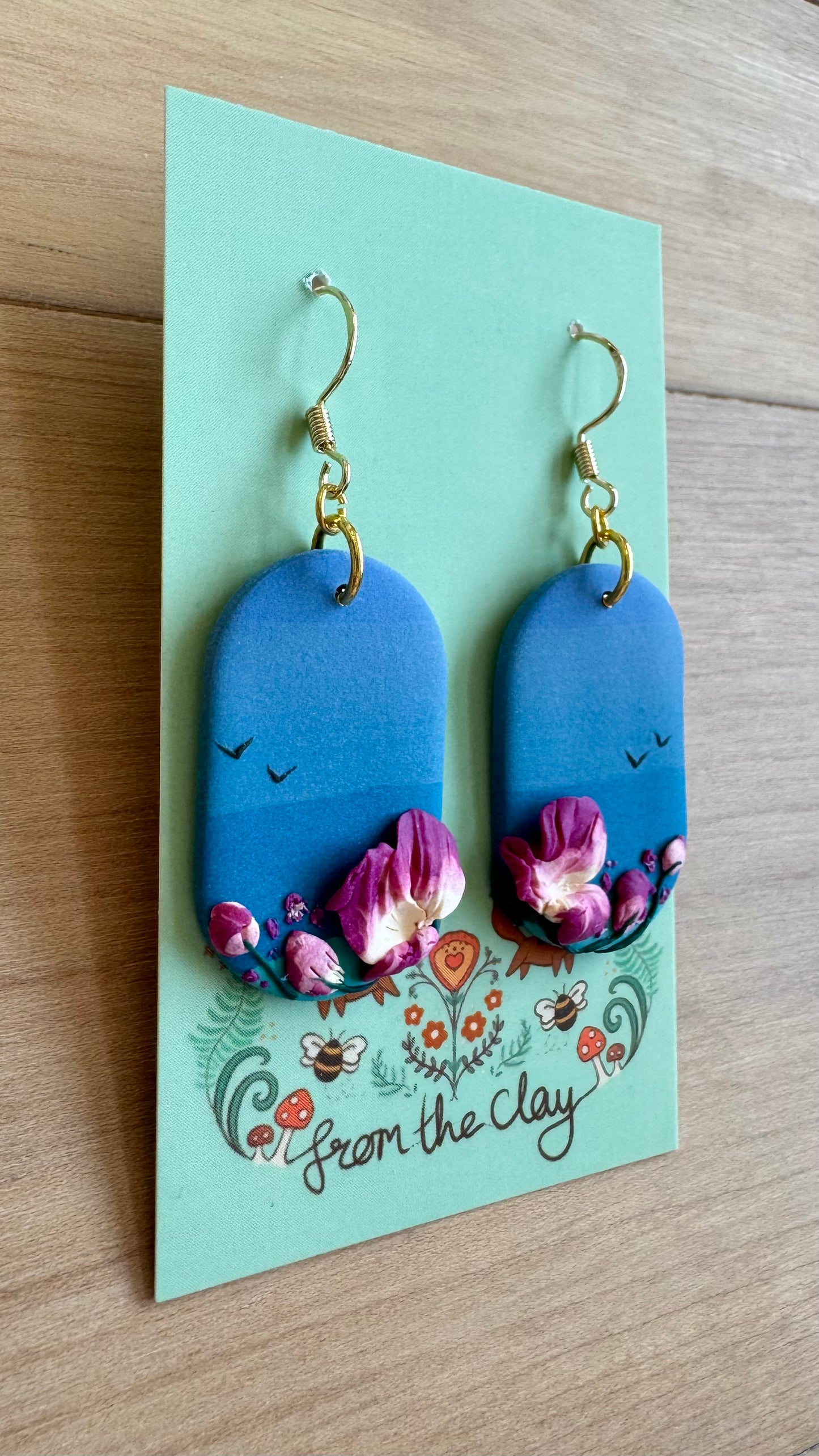 Blue earrings with floral design on a 'From the Clay' branded card, placed on a wooden surface.