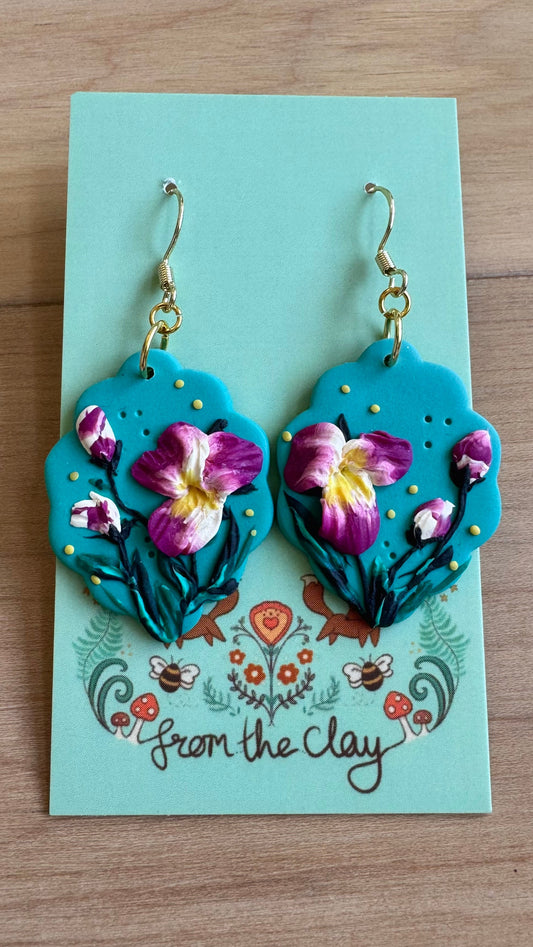 Earrings that have Purple & white Flowers on a blue background with a scalloped edge.  Placed on a 'From the Clay' branded card against a wooden background