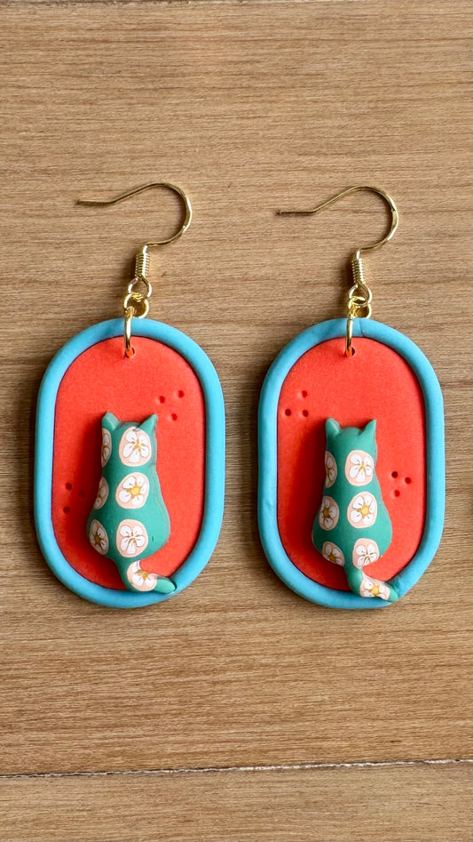 Pair of earrings with bright orange background and blue oval trim design featuring a puffy cat with a patterned surface.  