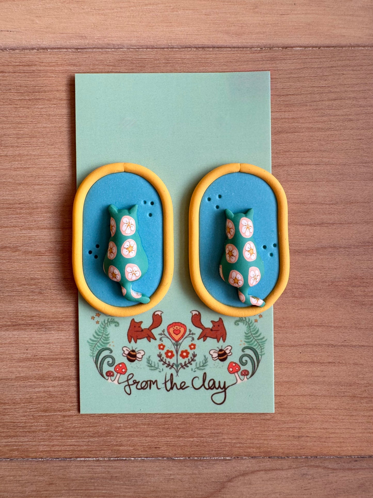 Oval blue earrings with Yellow trim, and patterned puffy cat in the middle.
Placed on a backing card on a wood surface 