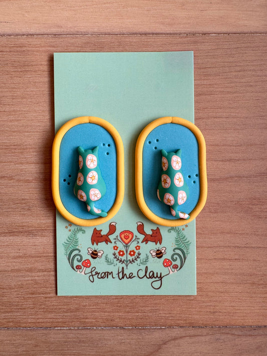 Oval blue earrings with Yellow trim, and patterned puffy cat in the middle.
Placed on a backing card on a wood surface 