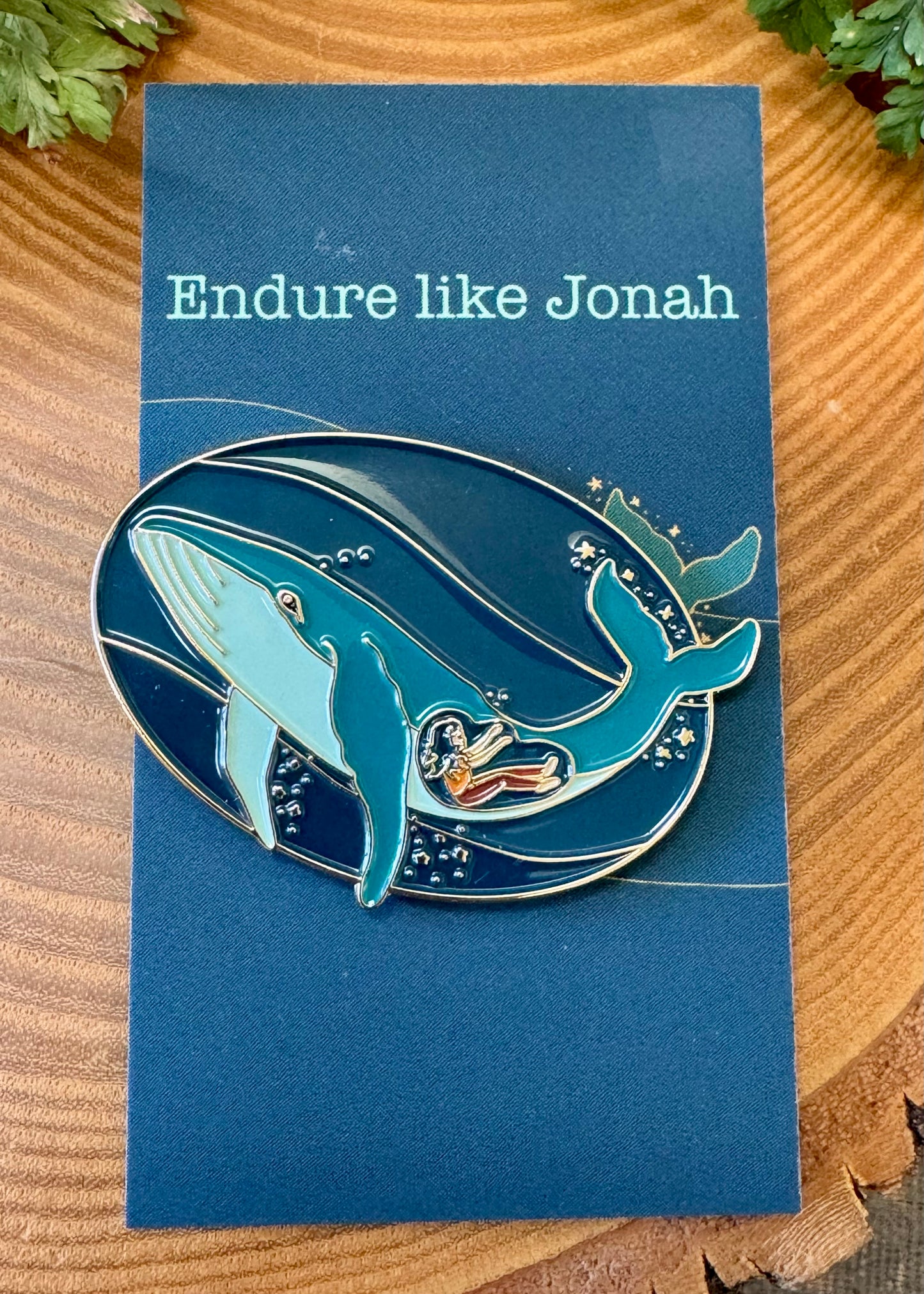 A pin and postcard set with an illustration of a whale and a person inside, titled 'Endure like Jonah'. The background is blue, and the text on the postcard reads the same as the illustration.
