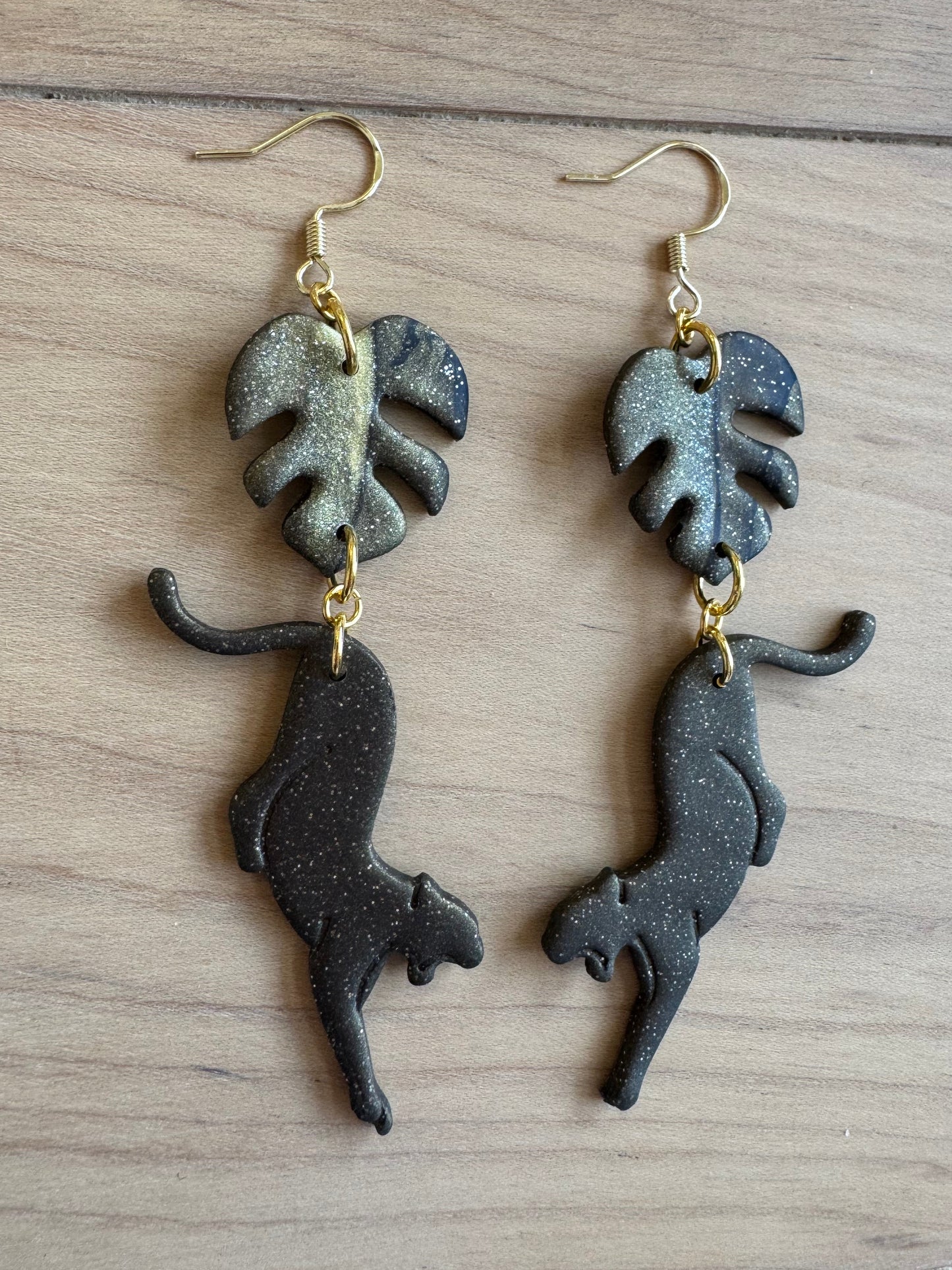 Pair of black and gold sparkly monstera leaf-shaped earrings with a black slightly sparkly panther attached. Placed  on a wooden surface