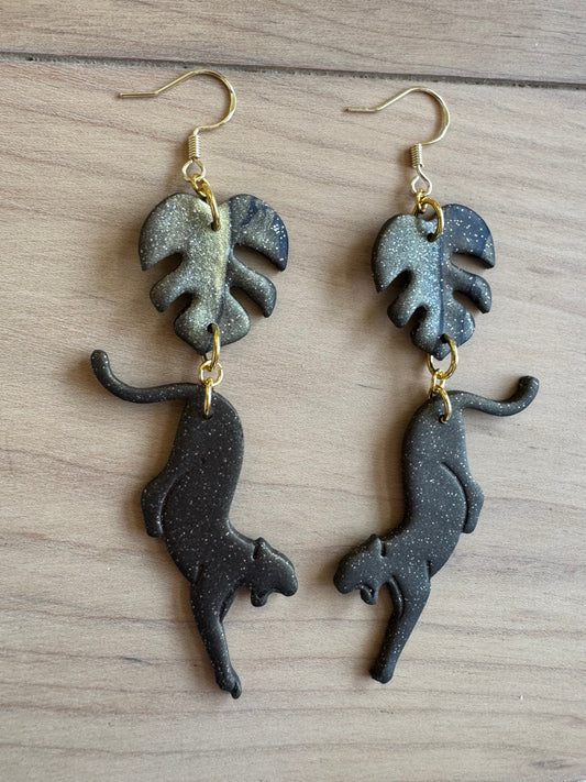 Pair of black and gold sparkly monstera leaf-shaped earrings with a black slightly sparkly panther attached. Placed  on a wooden surface