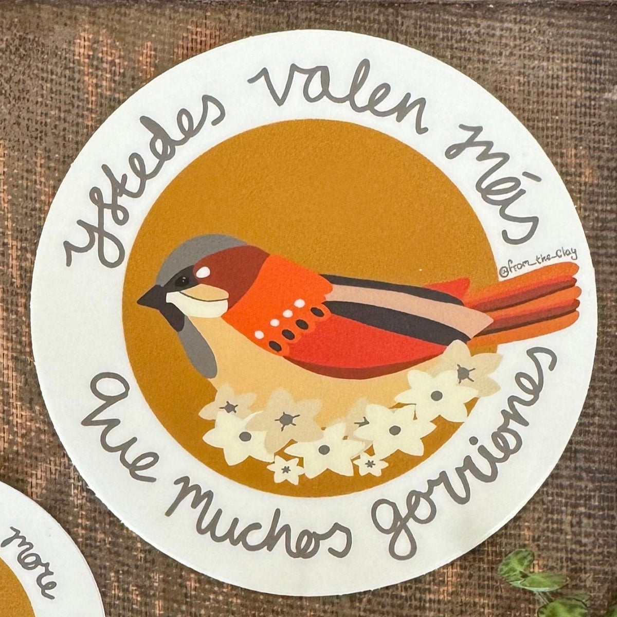 A circular waterproof sticker with an illustration of a sparrow sitting on cream colored flowers, with a mustard circular background, and the text 'you are worth more than many sparrows' but n Spanish so it says Ystedes valen mas que muchos gorriones around the outside in a cream border.
Based off of the scripture Matthew 10:31 
it is 2” across. 