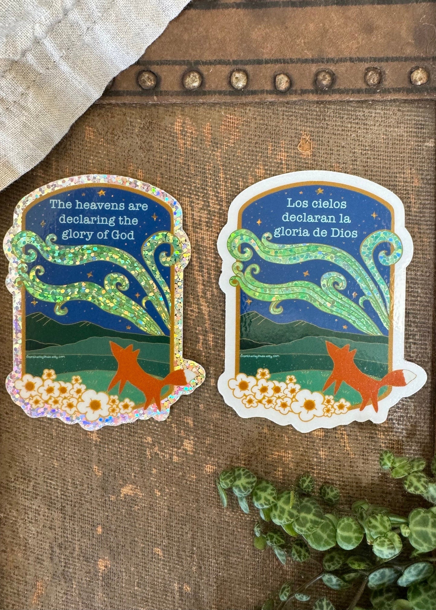 Two vinyl stickers featuring a fox looking at the Northern lights with a background of a clear sky at night. One sticker has the text 'The heavens are declareing the Glory of God' in English, and the other has the same text in Spanish.