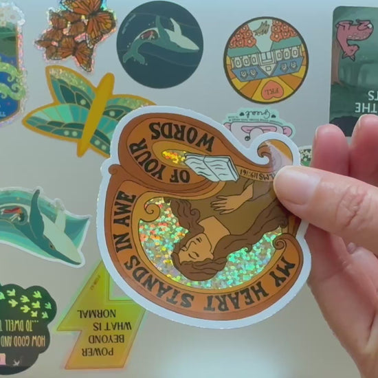 Video of the Sticker with a woman holding a book and text 'My heart stands in awe of your words' the sticker is being placed onto a laptop showing scale amongst others stickers on the laptop