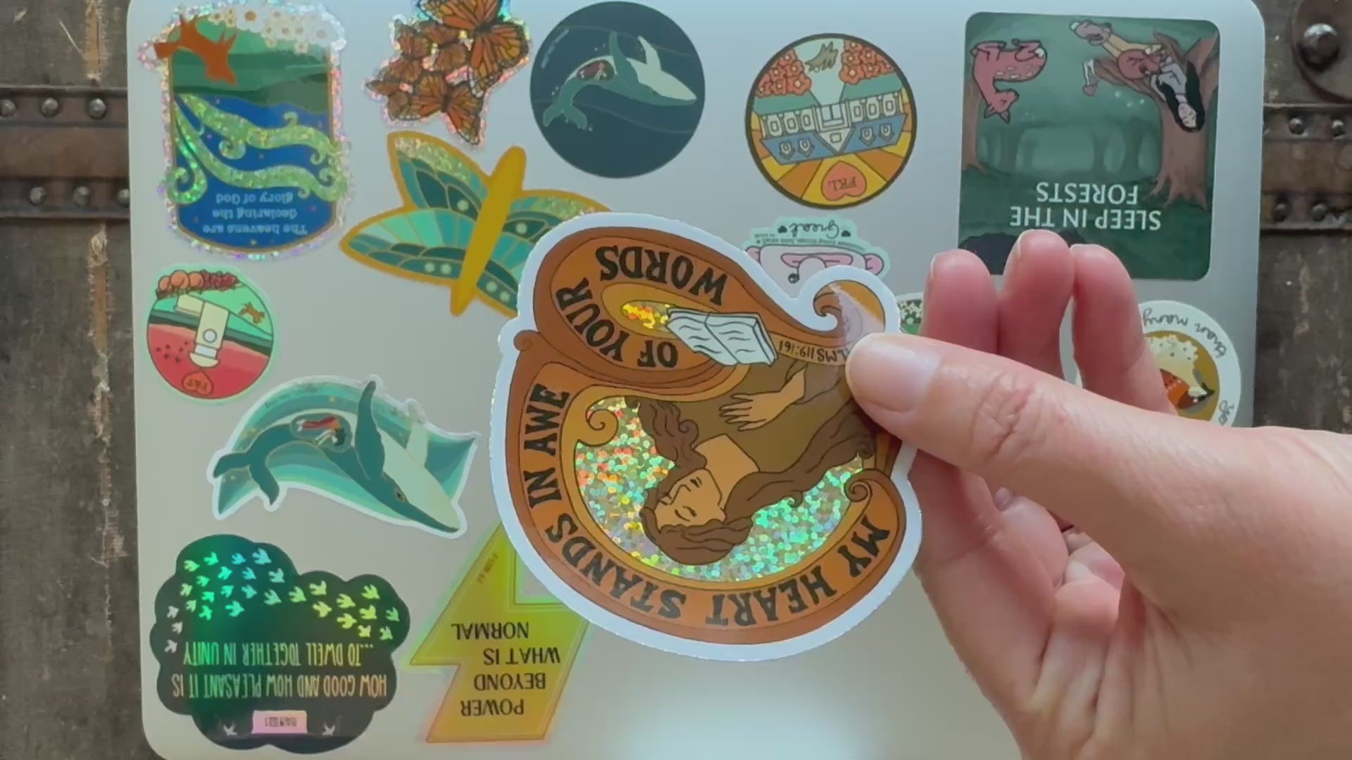 Video of the Sticker with a woman holding a book and text 'My heart stands in awe of your words' the sticker is being placed onto a laptop showing scale amongst others stickers on the laptop